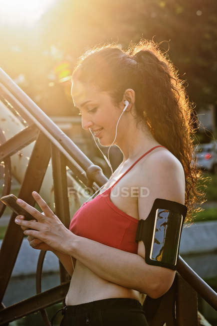Femme coureuse dans les écouteurs regardant smartphone dans la lumière du soleil — Photo de stock