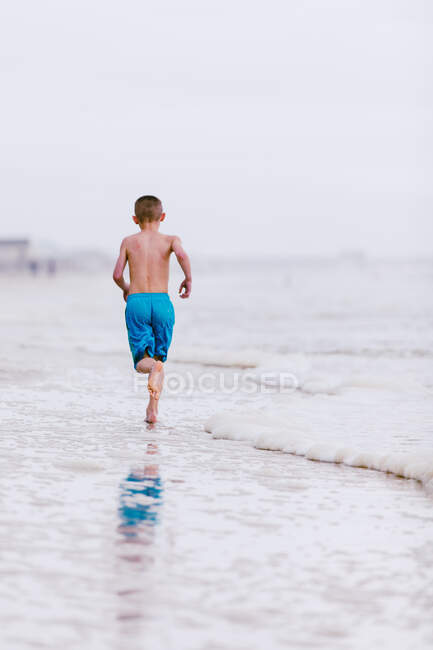 Хлопчик біжить вздовж краю води на пляжі, задній вид, острів Дофін, Алабама, США. — стокове фото