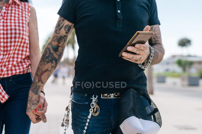 Пара татуированных хипстеров, держащихся за руки и смартфон, средняя секция — стоковое фото