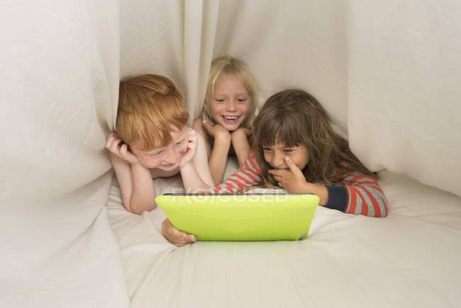 Дети лежат в постели с цифровым планшетом и смеются — стоковое фото