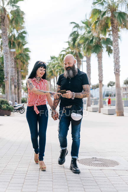 Mature hipster couple strolling while looking at smartphone on sidewalk, Valencia, Spain — Stock Photo