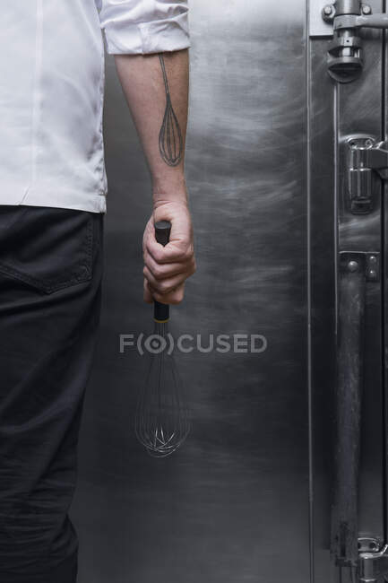 Cropped rear view shot of pastry chef with whisk tattoo holding whisk in kitchen — Stock Photo