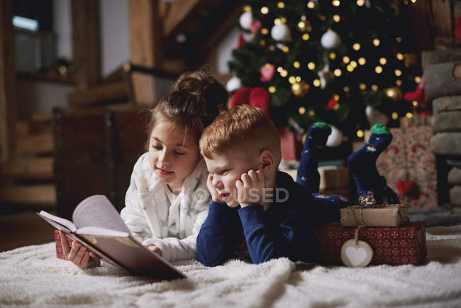 Young girl and boy reading book beside Christmas tree — Stock Photo