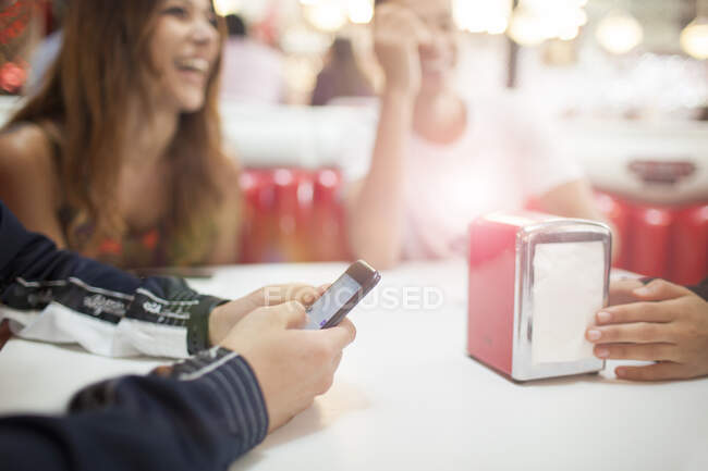 Grupo de jóvenes amigos sentados en la cafetería, hombre joven con teléfono inteligente, sección media - foto de stock
