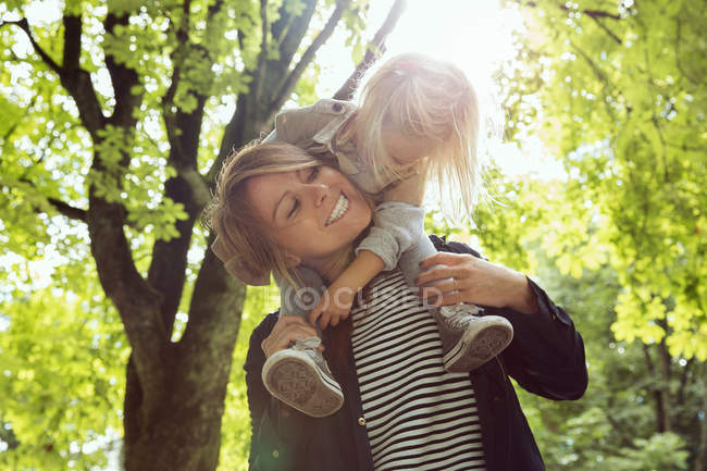 Mother giving toddler daughter on shoulders in sunlit park — Stock Photo