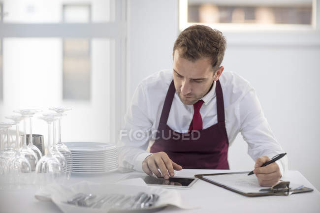Vista frontal de camarero en restaurante usando tableta digital y escritura en papel - foto de stock