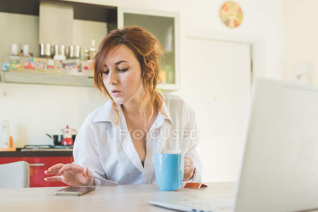 Молодая женщина за кухонным столом смотрит на смартфон — стоковое фото