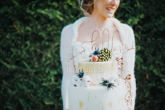Braut hält Hochzeitstorte mit Hecke im Hintergrund — Stockfoto