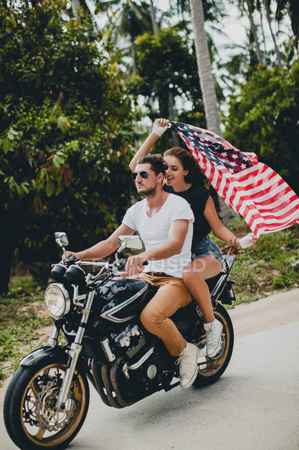 Pareja joven que sostiene la bandera estadounidense mientras conduce una motocicleta en la carretera rural, Krabi, Tailandia - foto de stock