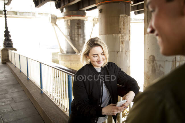 Jovem casal, de pé debaixo da ponte, jovem mulher usando smartphone — Fotografia de Stock