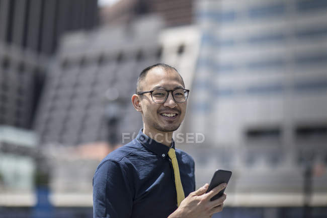 Retrato del hombre de negocios sonriente con teléfono inteligente fuera del edificio de oficinas - foto de stock