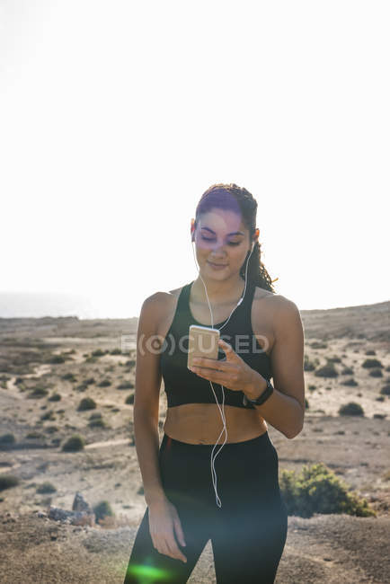 Young female runner looking at smartphone in arid coastal landscape — Stock Photo