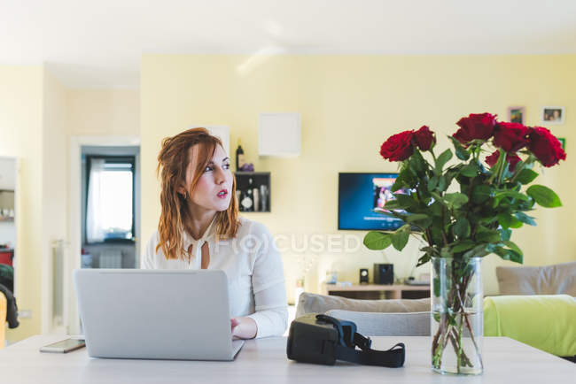 Молода жінка за столом у вітальні дивиться вбік, використовуючи ноутбук — стокове фото