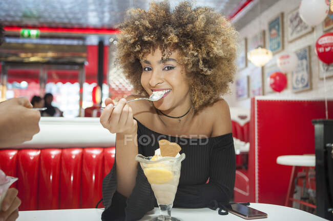 Retrato de una joven sentada en la cafetería, comiendo postre helado, sonriendo - foto de stock