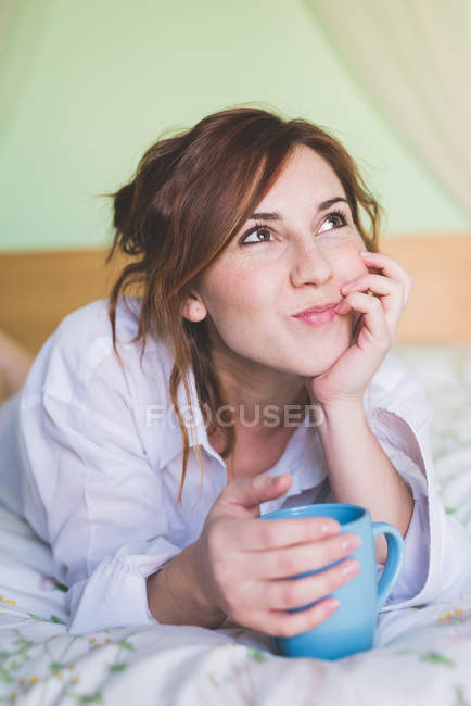 Junge Frau mit Kaffeetasse auf dem Bett und blickt auf — Stockfoto