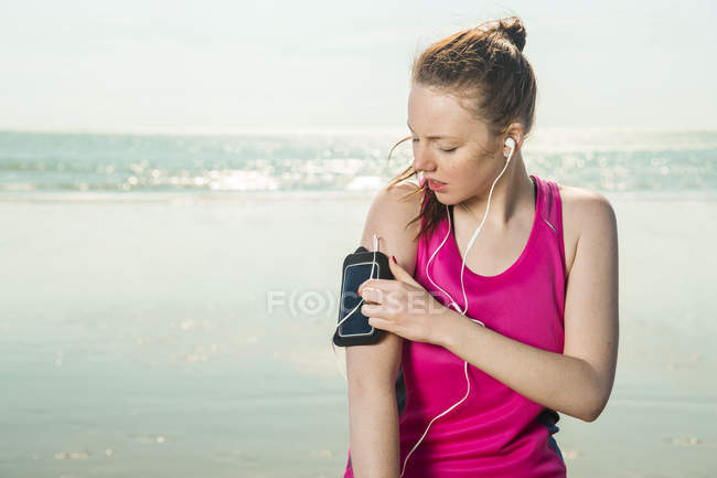 Mujer joven en auriculares ajustando la música en el brazalete en la playa - foto de stock