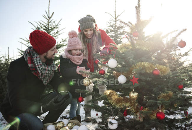 Chica y padres mirando bolas en el árbol de navidad del bosque - foto de stock
