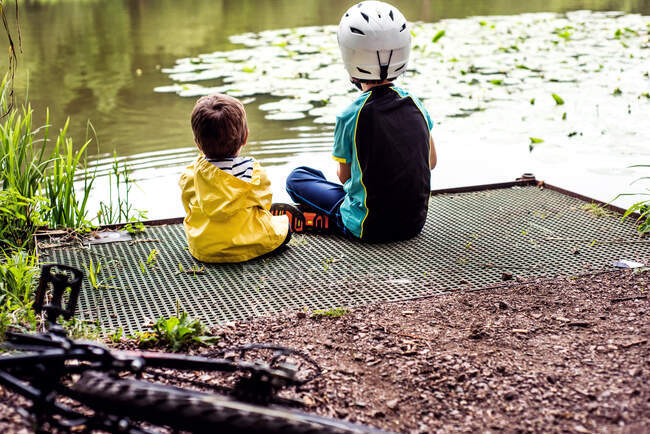 Dos hermanos jóvenes sentados en el borde del agua, vista trasera - foto de stock