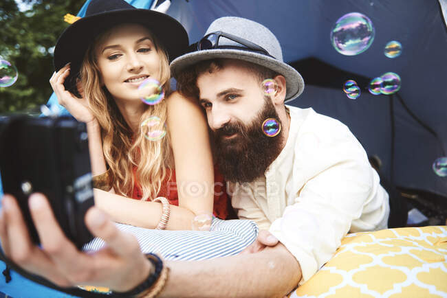 Young boho couple lying in tent taking selfie at festival — Stock Photo