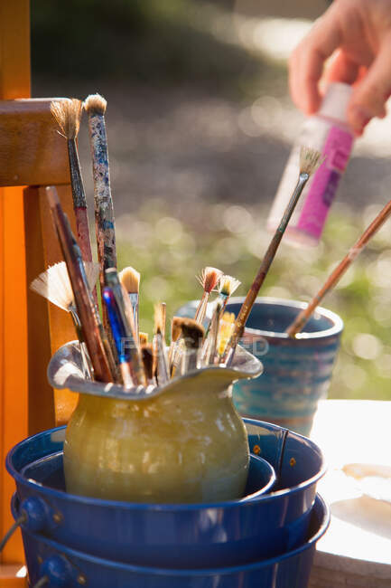 Pinceles en jarra y niña seleccionando pintura en el jardín, primer plano de la mano - foto de stock