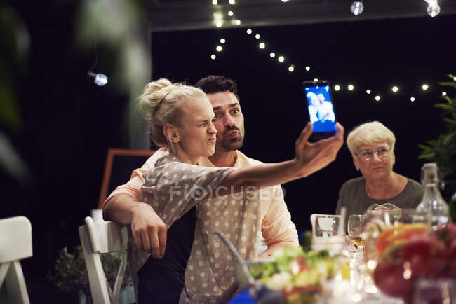 Група людей, які сидять за столом, насолоджуються їжею, молода жінка приймає селфі за допомогою смартфона — стокове фото