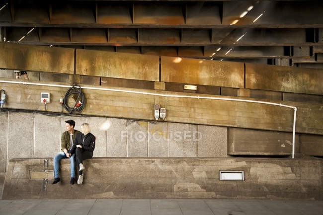 Jovem casal sentado no banco debaixo da ponte — Fotografia de Stock