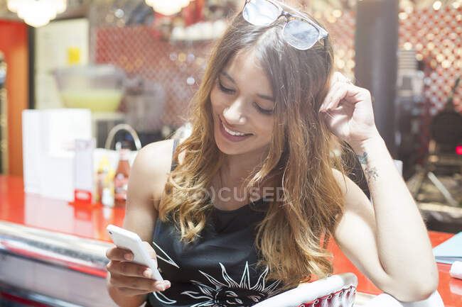 Junge Frau sitzt im Café, schaut aufs Smartphone, lächelt — Stockfoto