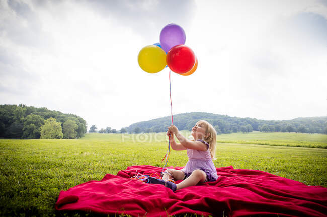 Girl sitting on red blanket in rural field looking up at bunch of colourful balloons — Stock Photo