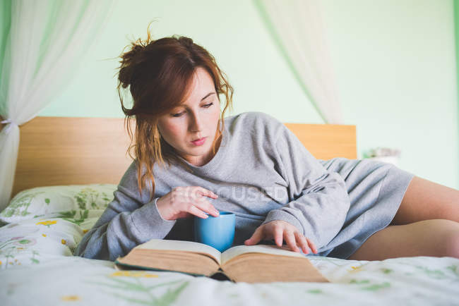 Молода жінка сидить на ліжку читаючи книгу — стокове фото