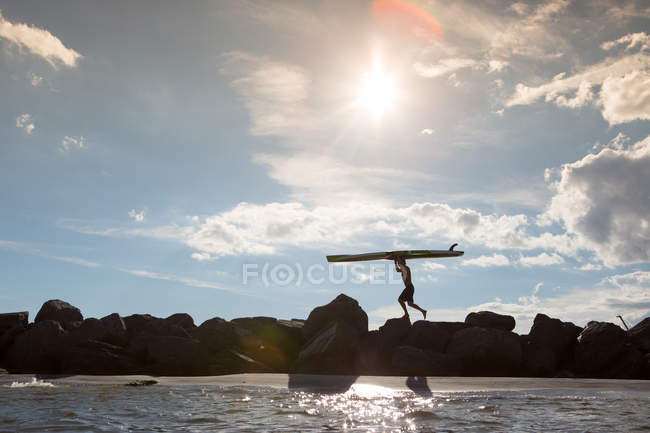 Vue lointaine de l'homme portant une planche de surf sur des rochers — Photo de stock