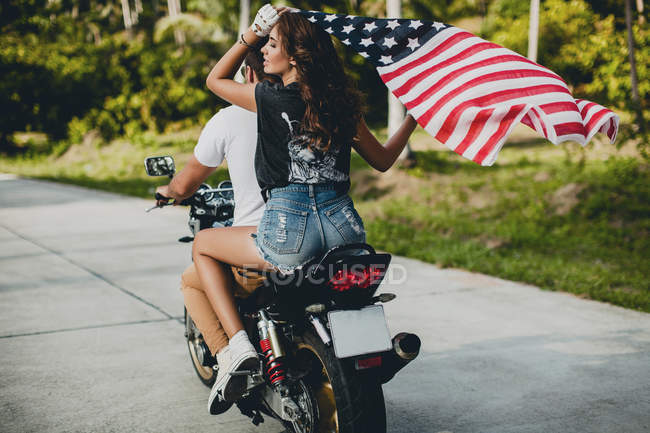Pareja joven sosteniendo la bandera estadounidense mientras conduce una motocicleta en la carretera rural, Krabi, Tailandia, vista trasera - foto de stock