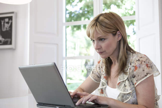 Mulher madura digitando no laptop por janela — Fotografia de Stock