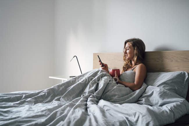 Jovem mulher sentada na cama com smartphone — Fotografia de Stock