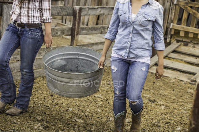 Deux jeunes femmes travaillant à la ferme, portant un seau en métal, section basse — Photo de stock