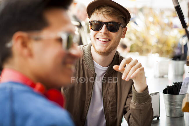 Dos amigos de pie en la cafetería, al aire libre, sonriendo - foto de stock