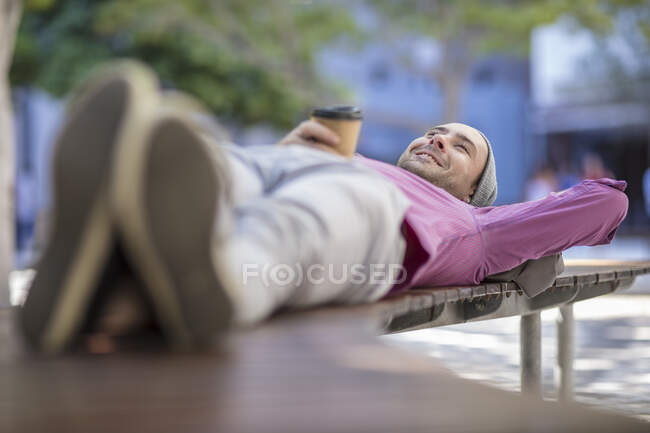 Jovem ao ar livre, deitado no banco, segurando taça de café takeaway — Fotografia de Stock