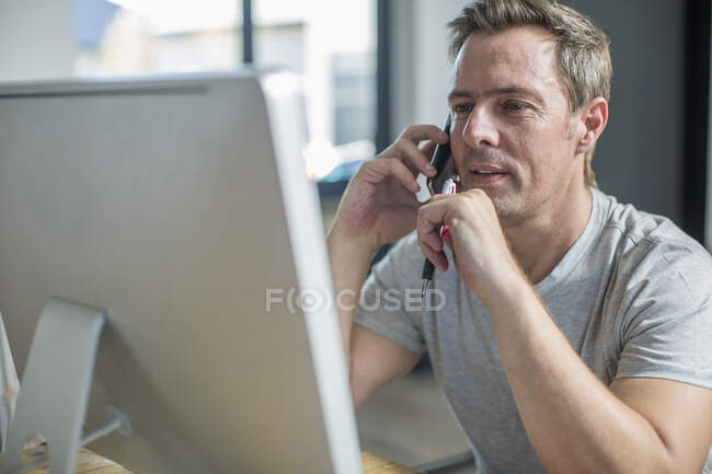 Hombre en el ordenador usando el teléfono inteligente para hacer una llamada telefónica - foto de stock