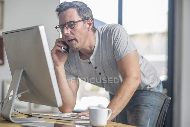 Man at computer using smartphone to make telephone call — Stock Photo