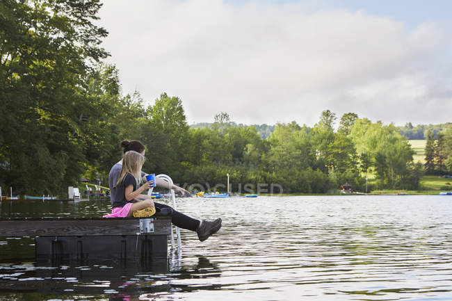 Two girls sitting on jetty and looking into lake — Stock Photo