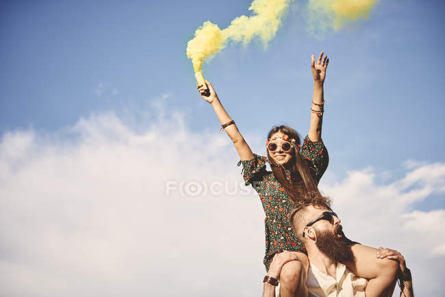 Young boho woman holding yellow smoke flare on boyfriend shoulders at festival — Stock Photo