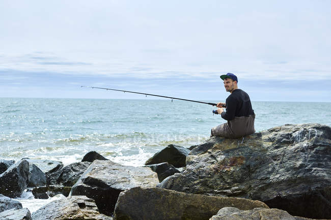 Jeune homme assis sur la mer de roche pêche — Photo de stock