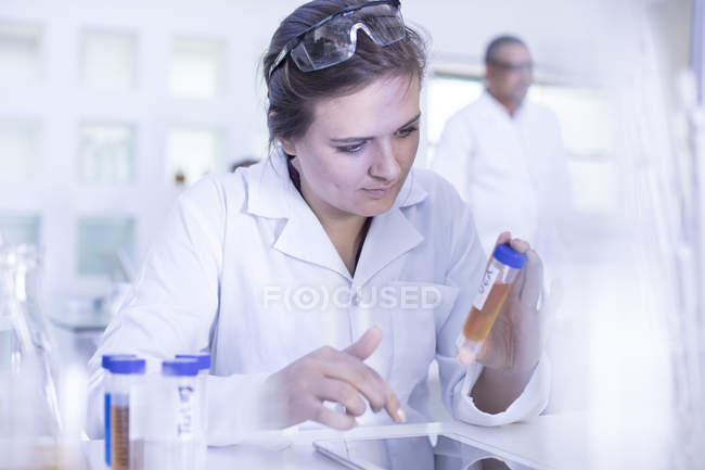Trabajador de laboratorio sosteniendo tubo de ensayo lleno de líquido, utilizando tableta digital - foto de stock