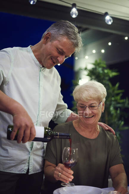 Senior mulher sentada à mesa de jantar, segurando copo de vinho, homem maduro derramando vinho em vidro — Fotografia de Stock