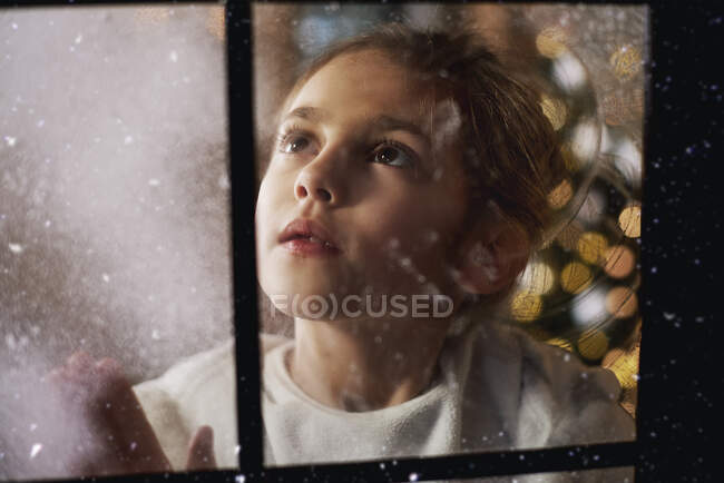 Giovane ragazza che guarda fuori dalla finestra, albero di Natale sullo sfondo dietro di lei, visto attraverso la finestra — Foto stock