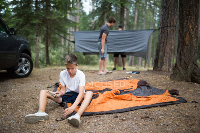 Adolescente menino martelando no tenda peg, amigos no fundo — Fotografia de Stock