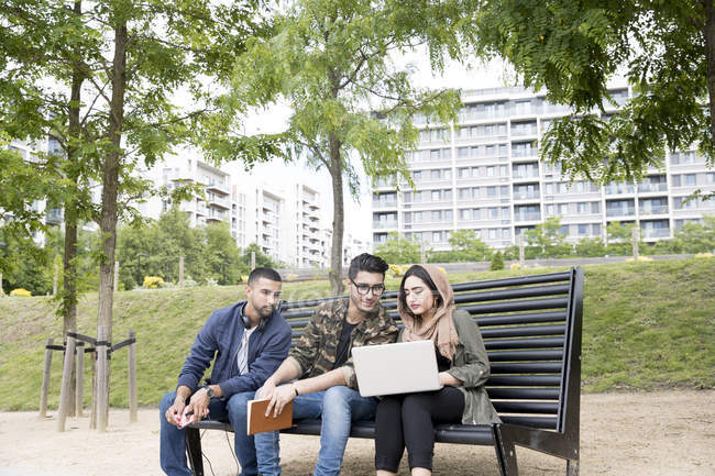 Tres amigos sentados en el banco en el parque y mirando el portátil - foto de stock