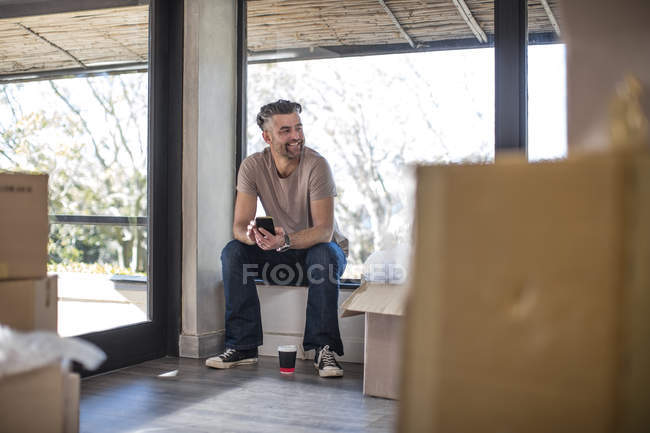 Uomo seduto in casa non arredata circondata da scatole di cartone e utilizzando smartphone — Foto stock