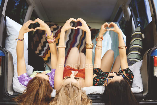 Trois jeunes femmes allongées sur le dos dans une botte de voiture faisant signe de coeur — Photo de stock