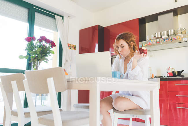 Молода жінка за кухонним столом дивиться на ноутбук і їсть яблуко — стокове фото