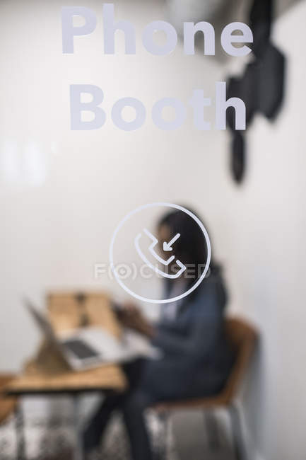 Empresária sentada na cabine telefônica do escritório, usando laptop, segurando smartphone, foco diferencial — Fotografia de Stock